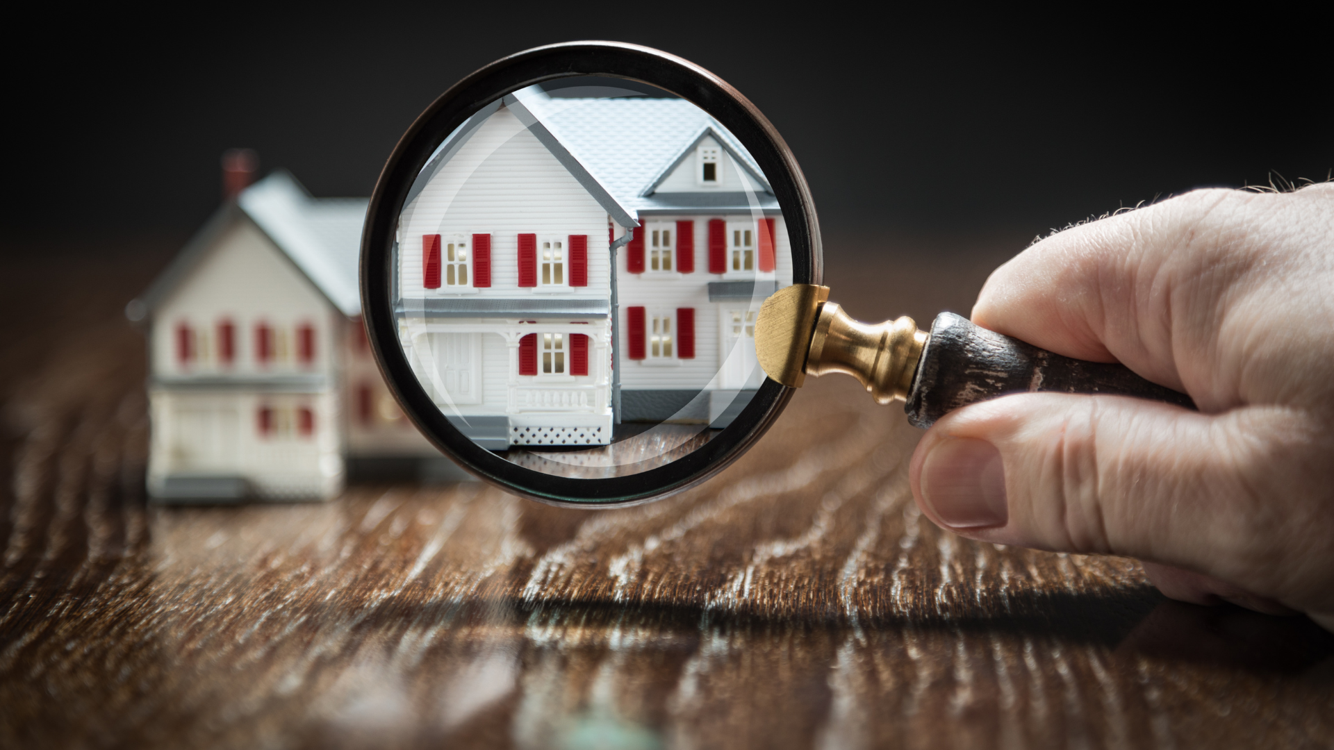 A person using a magnifying glass to examine a model house and mortgage documents, representing the scrutiny and understanding required for mortgage product transfers.